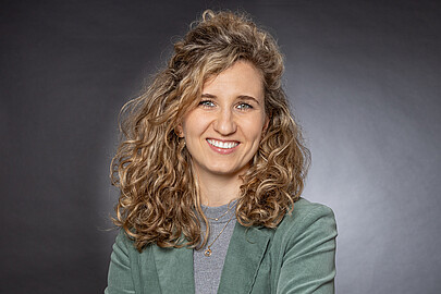 weiße Frau mit halblangem, lockigem, blonden Haar und mint-grünem Jacket lächelt in die Kamera