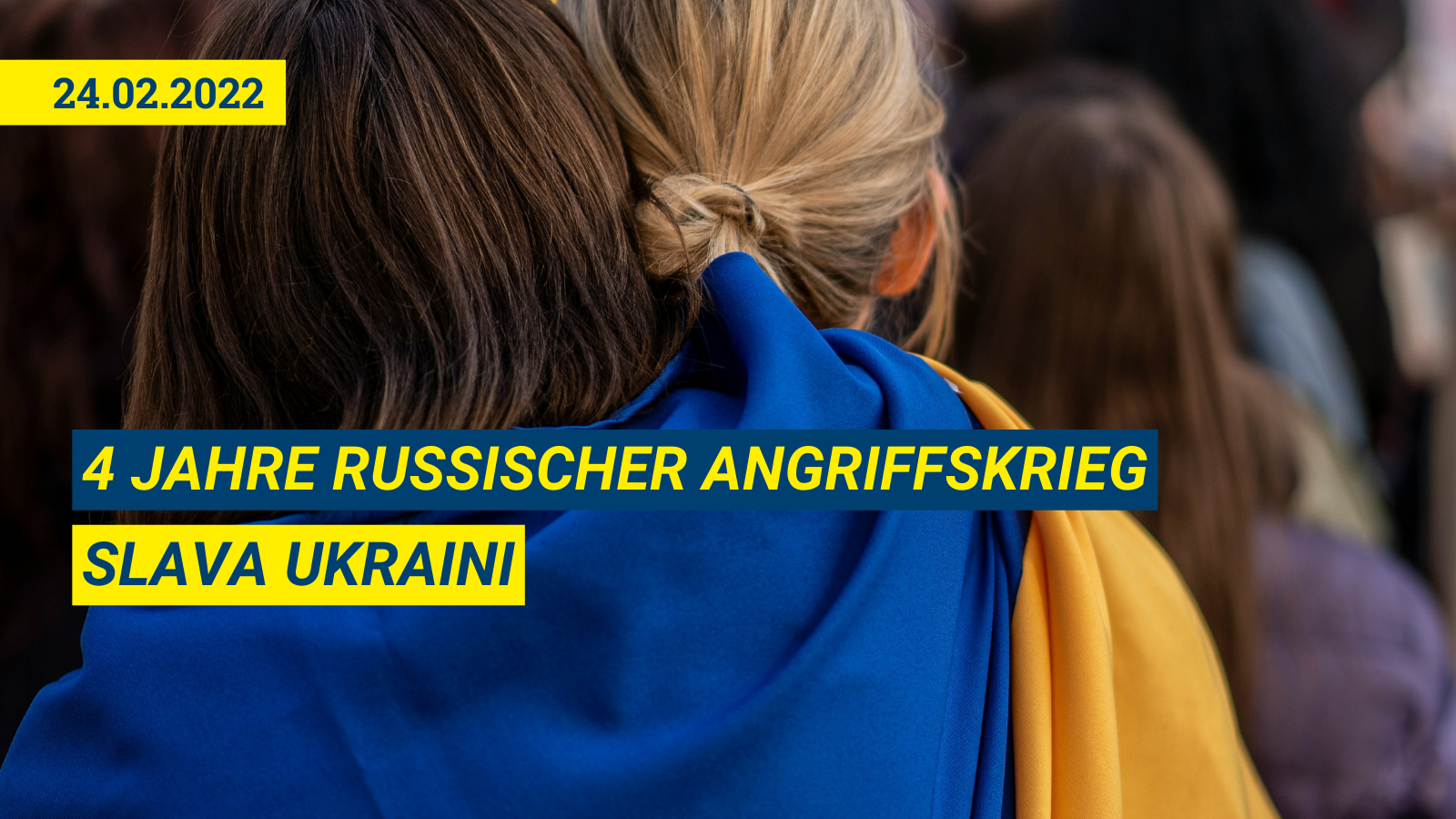 Nahaufnahme von zwei Personen von hinten, die eng umschlungen beieinander stehen. Sie tragen eine große ukrainische Flagge (blau und gelb) wie einen gemeinsamen Umhang über ihren Schultern. Im unscharfen Hintergrund sind weitere Menschen und Flaggen einer Demonstration zu sehen.