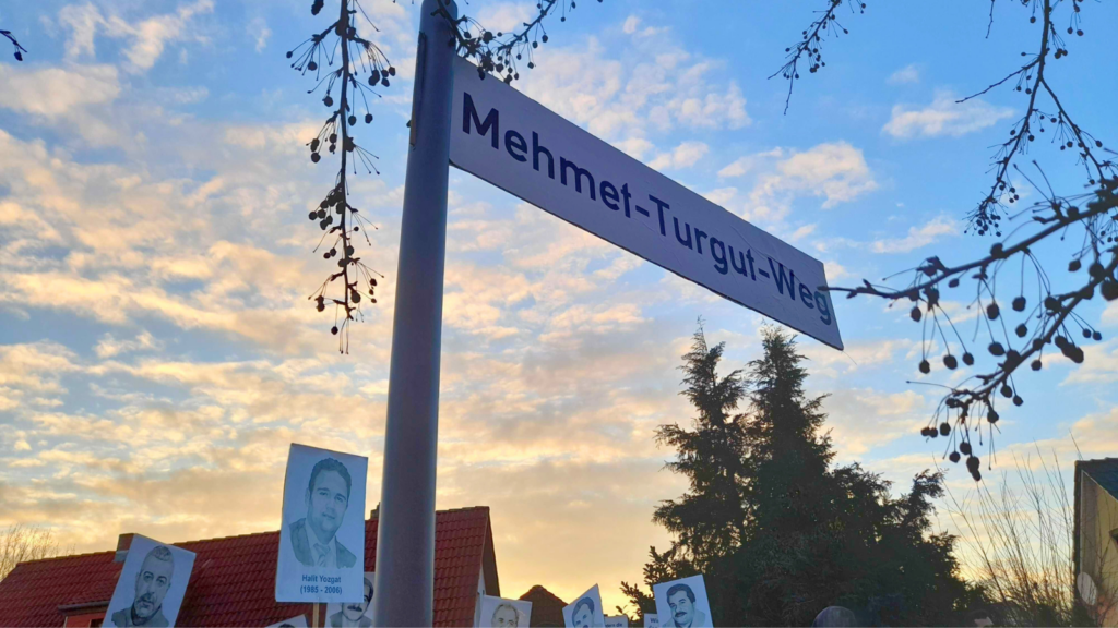 Straßenschild mit der Aufschrift „Mehmet-Turgut-Weg“ vor Abendhimmel. Darunter halten Menschen Porträts von NSU-Opfern bei einer Gedenkveranstaltung hoch.